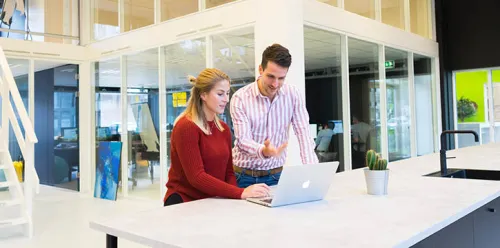 Deux collègues échangent devant un ordinateur dans un bureau moderne, symbolisant l'accompagnement Sofrecom.