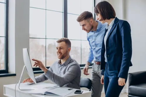 Trois professionnels analysent un projet sur ordinateur dans un bureau moderne, illustrant le travail collaboratif et l'expertise en transformation réseau.